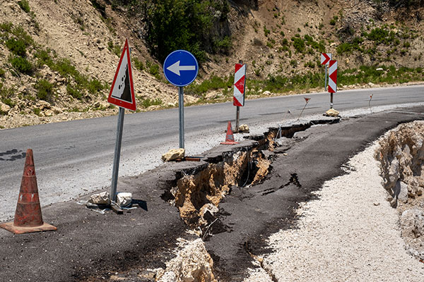 Image: The asphalt road has slipped with the soil under it. There are warning signs on the road. There is a big rift in the asphalt.