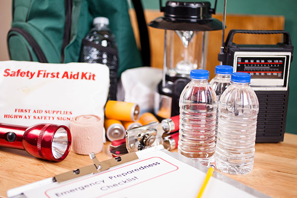 Image: Emergency preparedness items on a table, including a safety first aid kit, bottles of water, a flashlight, a lantern, a portable radio, bandages, batteries, and an emergency preparedness checklist